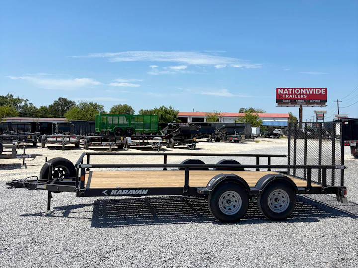 Driver-side profile of the black Karavan tandem-axle utility trailer with wood deck, mesh ramp gate, side rails, and spare tire.