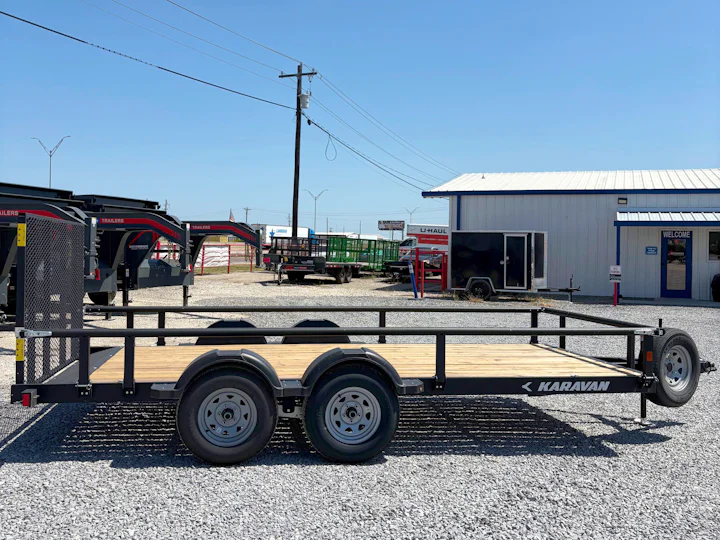 Passenger-side profile of the black Karavan tandem-axle utility trailer with wood deck, mesh ramp gate, side rails, and spare tire.